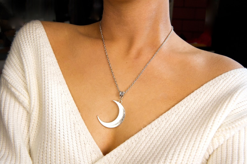 May include: A silver crescent moon pendant necklace on a silver chain. The necklace is worn by a person with a white sweater.