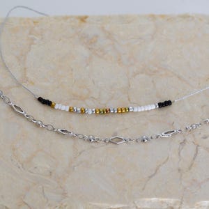 Pode incluir: Dois colares, uma corrente de prata com elos em forma de diamante e um colar de contas pretas, brancas e douradas.