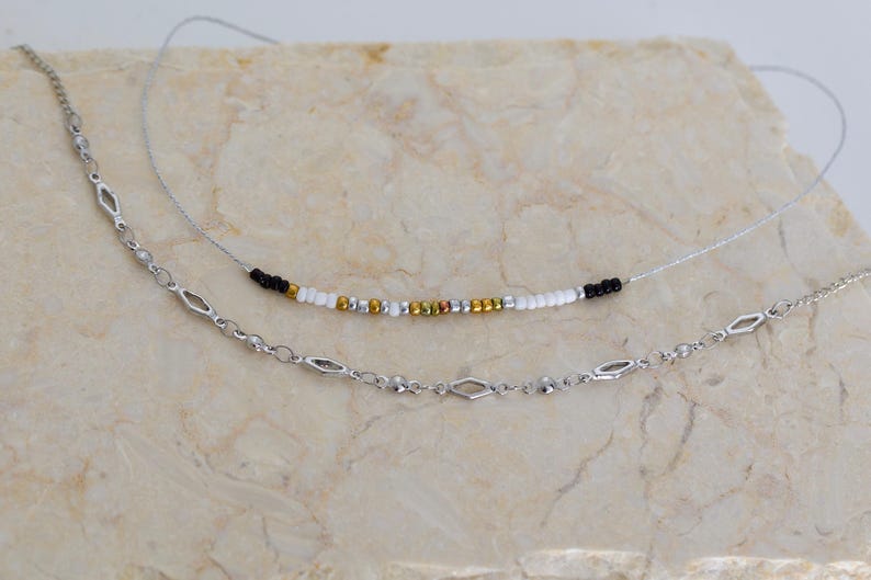 Pode incluir: Dois colares, uma corrente de prata com elos em forma de diamante e um colar de contas pretas, brancas e douradas.