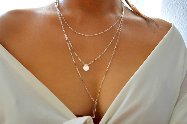 May include: A silver layered necklace with a round pendant and a long, thin bar pendant.