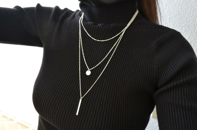 May include: A layered silver necklace with a round pendant and a long, thin bar pendant.