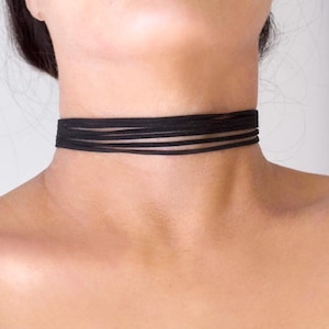 Puede incluir: Un collar gargantilla negro con múltiples bandas finas y superpuestas. La gargantilla se coloca alrededor del cuello, mostrando un diseño minimalista y versátil. El accesorio es de color oscuro y sólido.
