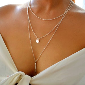 May include: A silver layered necklace with a round pendant and a long, thin bar pendant.