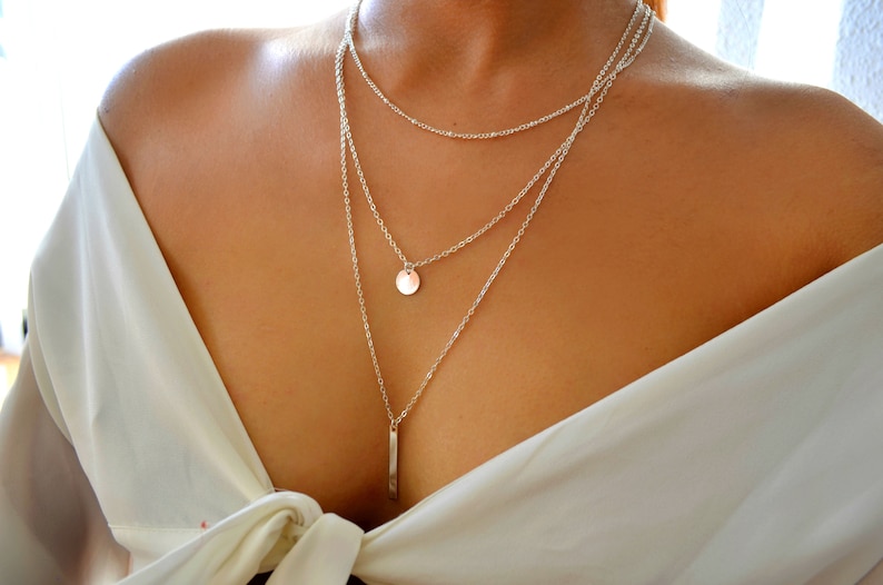 May include: A silver layered necklace with a round pendant and a long, thin bar pendant.