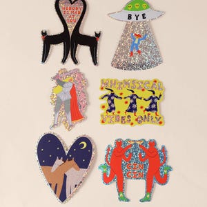 K&ouml;nnte beinhalten: Eine Sammlung von sechs holografischen Aufklebern mit verschiedenen Motiven: zwei schwarze Katzen in Herzform mit dem Text NOBODY IS MAD AT YOU, ein UFO mit dem Text BYE, ein Ritter, drei Figuren mit dem Text WHIMSICAL VIBES ONLY, zwei Pferde in einem Herz und zwei rote teufelsartige Figuren mit dem Text CIN CIN.