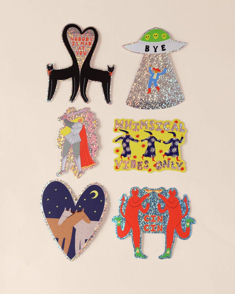 K&ouml;nnte beinhalten: Eine Sammlung von sechs holografischen Aufklebern mit verschiedenen Motiven: zwei schwarze Katzen in Herzform mit dem Text NOBODY IS MAD AT YOU, ein UFO mit dem Text BYE, ein Ritter, drei Figuren mit dem Text WHIMSICAL VIBES ONLY, zwei Pferde in einem Herz und zwei rote teufelsartige Figuren mit dem Text CIN CIN.