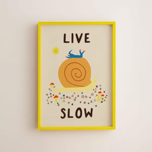 May include: A framed wall art print featuring a large yellow snail with a brown shell, a blue figure lying on top, and small red mushrooms and flowers below. The text LIVE SLOW is printed in dark brown letters above and below the illustration.