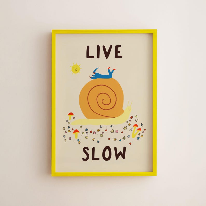 May include: A framed wall art print featuring a large yellow snail with a brown shell, a blue figure lying on top, and small red mushrooms and flowers below. The text LIVE SLOW is printed in dark brown letters above and below the illustration.