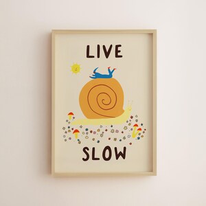 May include: A framed wall art print on a cream background featuring a large yellow snail with an orange shell. A blue figure lies on the shell. Below the snail are small colorful flowers and mushrooms. The text reads LIVE at the top and SLOW at the bottom.