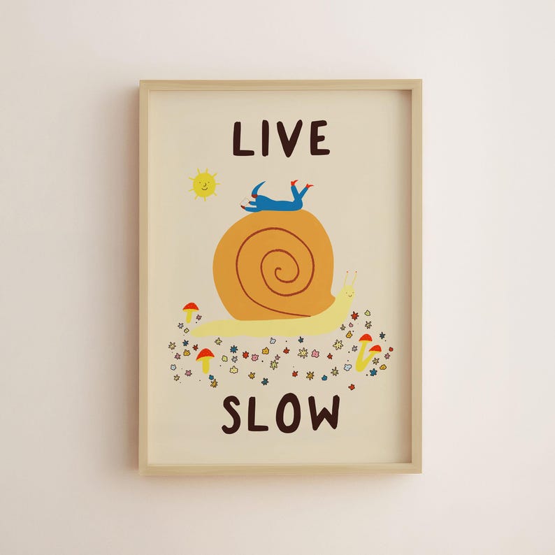 May include: A framed wall art print on a cream background featuring a large yellow snail with an orange shell. A blue figure lies on the shell. Below the snail are small colorful flowers and mushrooms. The text reads LIVE at the top and SLOW at the bottom.