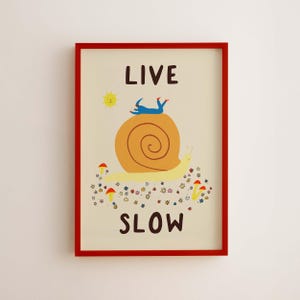 May include: A framed wall art print with a cream background featuring a large yellow snail with a brown shell. A blue figure lies on the shell. The text reads LIVE SLOW. There is a yellow sun and small red mushrooms and colorful flowers below.