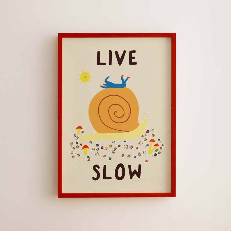May include: A framed wall art print with a cream background featuring a large yellow snail with a brown shell. A blue figure lies on the shell. The text reads LIVE SLOW. There is a yellow sun and small red mushrooms and colorful flowers below.