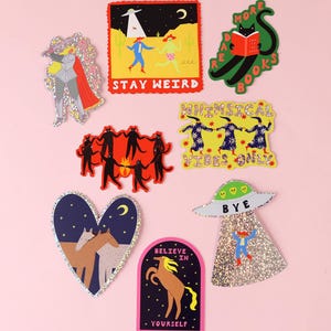 K&ouml;nnte beinhalten: Eine Sammlung von acht bunten Aufklebern mit verschiedenen Illustrationen, darunter ein Ritter, ein UFO, eine lesende schwarze Katze, tanzende Figuren, Pferde und Texte wie STAY WEIRD, MORE BOOKS, WHIMSICAL VIBES ONLY, BYE und BELIEVE IN YOURSELF.
