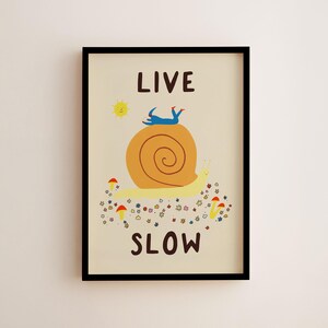 May include: A framed wall art print on a cream background featuring the text LIVE SLOW in dark brown. The illustration depicts a large yellow snail with an orange shell, a small blue figure lying on top, a yellow sun, and colorful flowers and mushrooms.