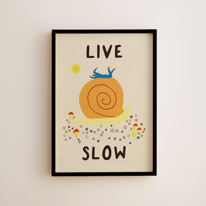 May include: A framed wall art print on a cream background featuring the text LIVE SLOW in dark brown. The illustration depicts a large yellow snail with an orange shell, a small blue figure lying on top, a yellow sun, and colorful flowers and mushrooms.