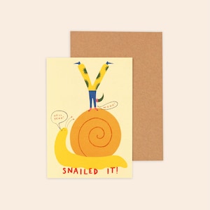 May include: A greeting card with a yellow snail and a person doing a handstand on its shell. The snail has a speech bubble saying HELL YEAH! and the person has one saying WOOO!. The text SNAILED IT! is printed at the bottom in red letters.