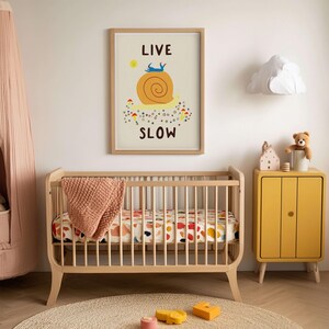 May include: A framed wall art print featuring a large yellow snail with a blue figure on its shell, a small yellow sun, and colorful mushrooms. The text reads LIVE SLOW in dark brown letters. The print is displayed in a light wood frame.