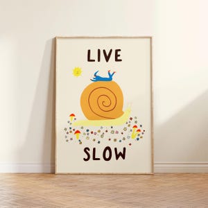 May include: A rectangular wall art print on a cream background featuring the text LIVE SLOW in dark brown. The illustration depicts a large yellow snail with an orange shell, a small blue figure lying on top, a yellow sun, and colorful flowers and mushrooms.