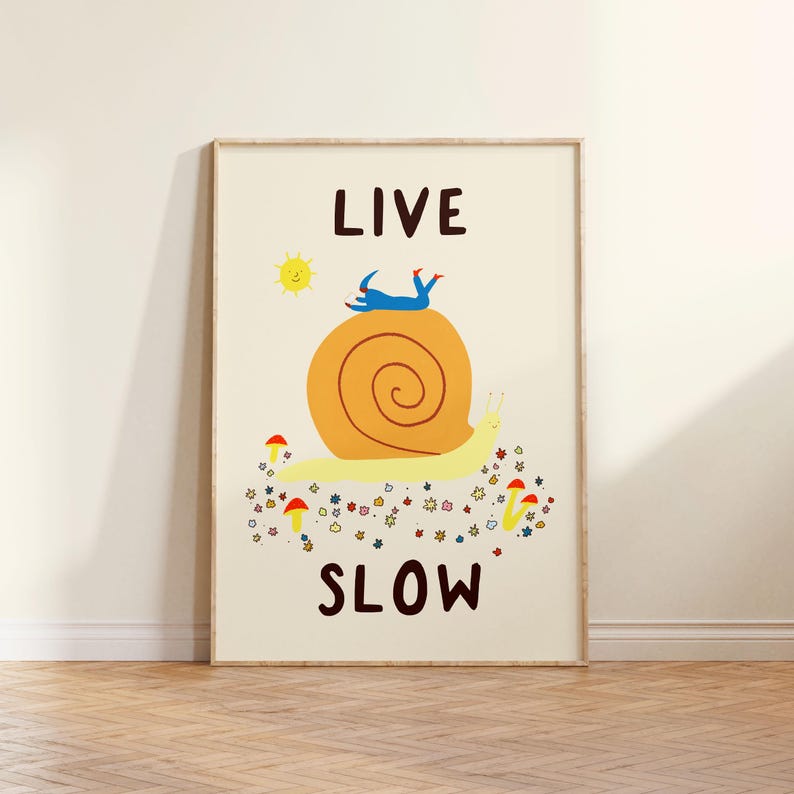 May include: A rectangular wall art print on a cream background featuring the text LIVE SLOW in dark brown. The illustration depicts a large yellow snail with an orange shell, a small blue figure lying on top, a yellow sun, and colorful flowers and mushrooms.