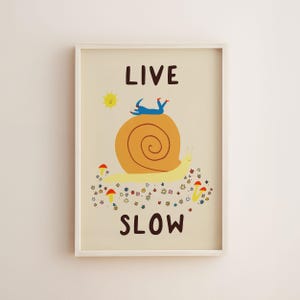 May include: A framed wall art print on a cream background featuring a large yellow snail with an orange shell. A blue figure lies on the shell. Below are small colorful flowers and red mushrooms. The text reads LIVE at the top and SLOW at the bottom.