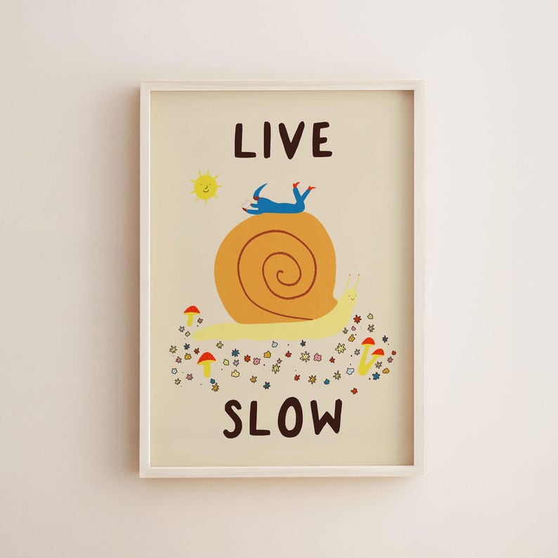 May include: A framed wall art print on a cream background featuring a large yellow snail with an orange shell. A blue figure lies on the shell. Below are small colorful flowers and red mushrooms. The text reads LIVE at the top and SLOW at the bottom.