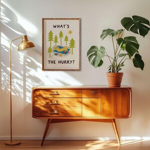May include: A rectangular wall art print in a wooden frame featuring a yellow figure lying on a blue blanket amidst green pine trees and a yellow sun. The text "WHAT'S THE HURRY?" is printed in black capital letters above and below the illustration.