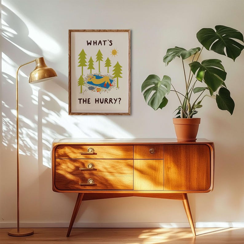 May include: A rectangular wall art print in a wooden frame featuring a yellow figure lying on a blue blanket amidst green pine trees and a yellow sun. The text "WHAT'S THE HURRY?" is printed in black capital letters above and below the illustration.