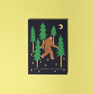 Op de afbeelding: Verjaardagskaart met een illustratie van een bruine Bigfoot die door een bos van groene bomen loopt onder een halve maan. De kaart heeft de tekst "HAPPY BIRTHDAY, YOU LEGEND!" met oranje paddenstoelen.