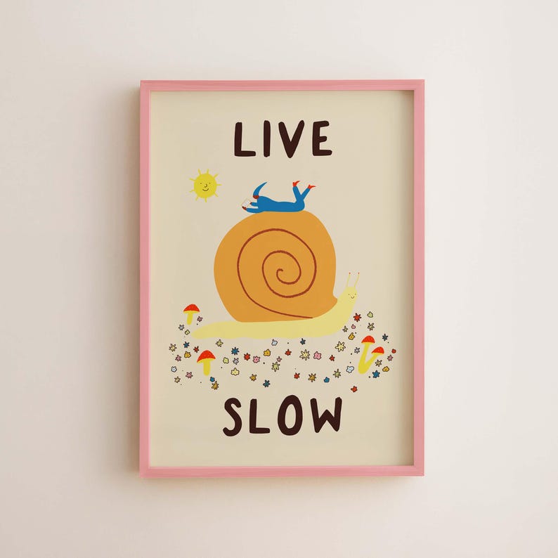 May include: A framed wall art print featuring a large yellow snail with a brown shell, a blue figure lying on top, and the text LIVE SLOW. The illustration includes a yellow sun, small red mushrooms, and colorful flowers on a cream background.