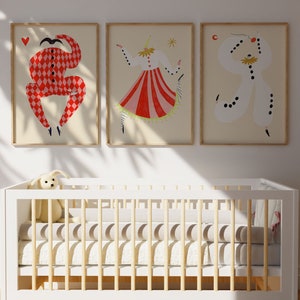May include: Three framed illustrations of stylized figures in red, white, and black costumes. The figures are in various poses, with one holding a heart, another holding a star, and the third holding a crescent moon. The illustrations are hung on a white wall above a white crib with a wooden frame.