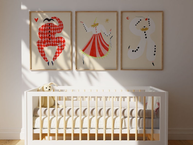 May include: Three framed illustrations of stylized figures in red, white, and black costumes. The figures are in various poses, with one holding a heart, another holding a star, and the third holding a crescent moon. The illustrations are hung on a white wall above a white crib with a wooden frame.