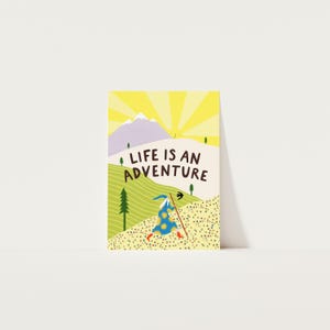 May include: A rectangular greeting card featuring a colorful illustration of a person in a blue sun-patterned robe and red boots walking with a staff. The scene includes green hills, purple mountains, a yellow sun, and the text LIFE IS AN ADVENTURE.