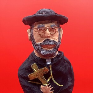 May include: A hand-painted figurine of St. Damien, a Catholic priest, holding a cross. The figurine is wearing a black robe and a black hat. The base of the figurine is white with the words "St. Damien" written in black.