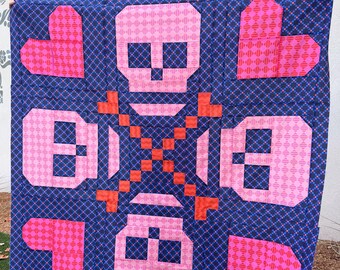 May include: A square quilt featuring a dark blue background with a repeating small pink diamond pattern. The design includes four bright pink hearts in the corners, four light pink skulls, and a central cross made of small orange square patches.