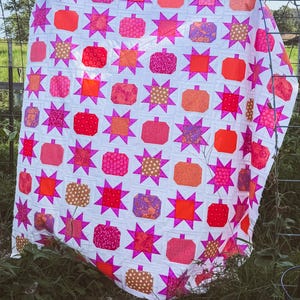 May include: A quilt featuring a repeating pattern of pumpkins and eight-pointed stars. The design uses a white background with various fabric squares in shades of bright pink, orange, red, and gold, showcasing diverse floral and geometric prints.