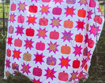May include: A quilt featuring a repeating pattern of pumpkins and eight-pointed stars. The design uses a white background with various fabric squares in shades of bright pink, orange, red, and gold, showcasing diverse floral and geometric prints.