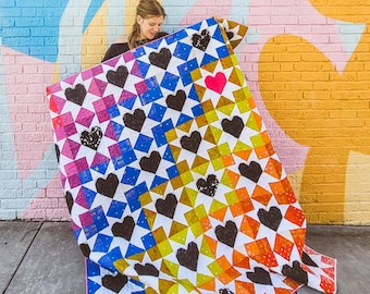 May include: A colorful quilt with a pattern of black hearts on a white background. The quilt is made up of squares of different colors, including blue, yellow, orange, and pink. The quilt is being held up by a person, and the background is a brick wall.
