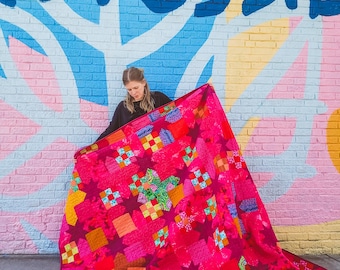 May include: A vibrant, handmade quilt in shades of pink, red, orange, and green. The quilt features a patchwork design with geometric shapes and star patterns. The background is a colorful brick wall with the words "IS POSSIBLE" in blue.