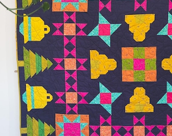 May include: A colorful quilt with a dark blue background, featuring repeating patterns of stars, bells, and Christmas trees in bright pink, orange, yellow, and green. The quilt is hanging on a wall.