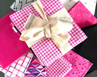 May include: A stack of assorted fabric squares featuring various patterns including pink gingham, magenta chevron, white with black stars and birds, and geometric shapes, all tied together with a beige fabric ribbon bow.