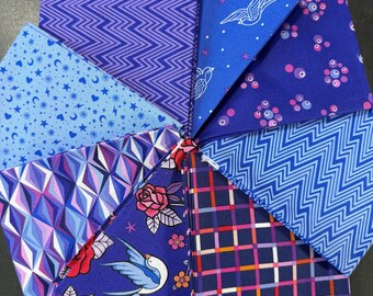 May include: A collection of eight square fabric swatches arranged in a circular fan pattern. The fabrics feature various blue and purple patterns, including zigzags, geometric shapes, plaid, floral motifs with birds, and small celestial symbols like stars.