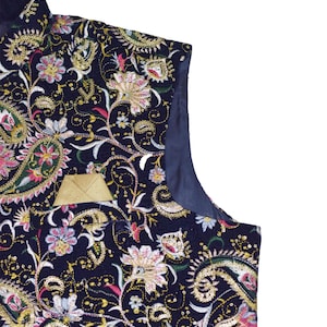 Op de afbeelding: Een marineblauwe vest met een ingewikkeld bloemen- en paisley patroon. Het ontwerp bevat gouden, roze en witte borduursels, met een klein gouden zakje. De vest heeft een satijnen voering en is een formeel kledingstuk.