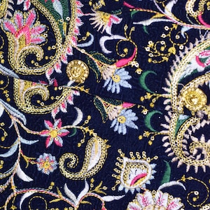Op de afbeelding: Close-up van een marineblauwe stof met ingewikkelde borduursels. Het ontwerp bevat paisley- en bloempatronen in goud, roze, wit en groen garen. De borduursels zijn gedetailleerd en sierlijk, wat een rijke, getextureerde uitstraling cre&euml;ert.