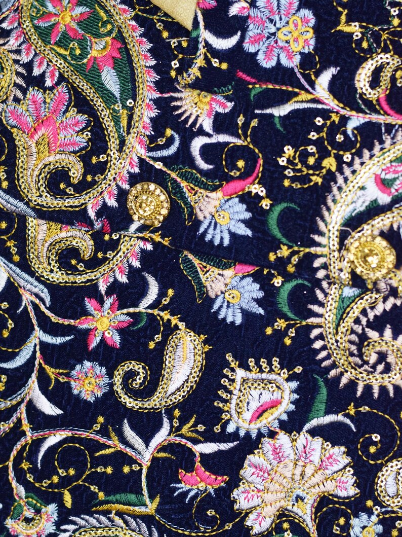 Op de afbeelding: Close-up van een marineblauwe stof met ingewikkelde borduursels. Het ontwerp bevat paisley- en bloempatronen in goud, roze, wit en groen garen. De borduursels zijn gedetailleerd en sierlijk, wat een rijke, getextureerde uitstraling cre&euml;ert.