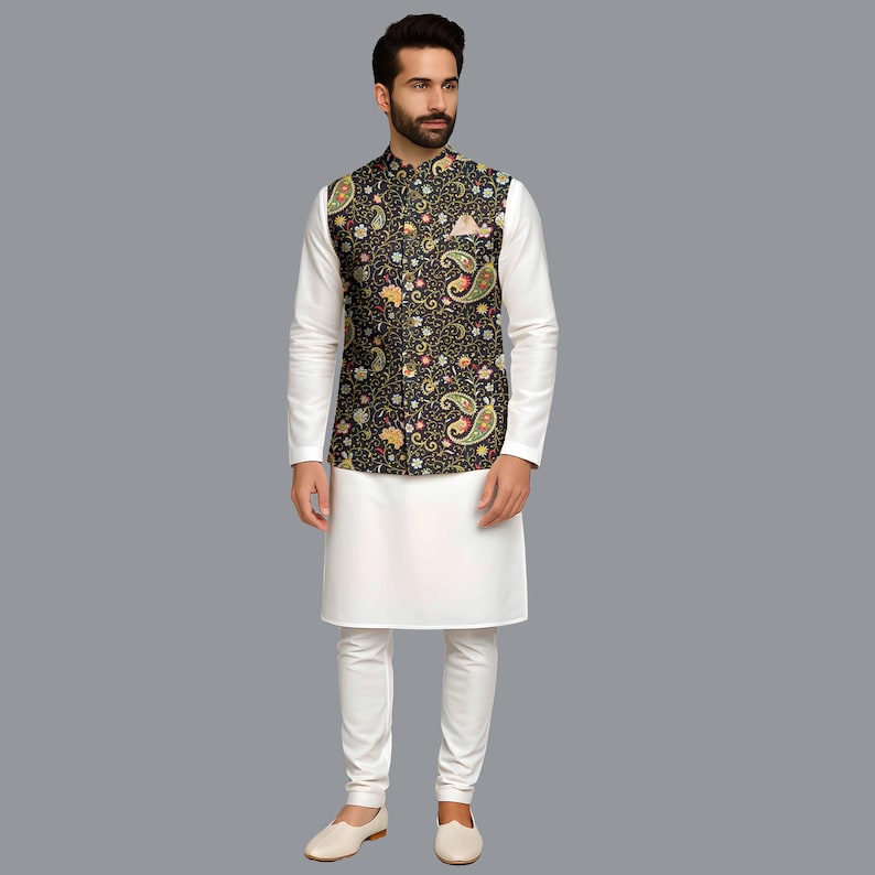 Op de afbeelding: Een zwart vest met paisley-patroon met een wit shirt met lange mouwen en een witte broek. Het vest heeft een kleurrijk bloemmotief met gele, rode en groene accenten. De outfit wordt gecompleteerd met witte schoenen.