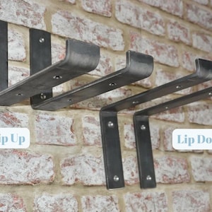 May include: Metal shelf brackets in a dark gray finish are mounted on a brick wall. The brackets are labeled "Lip Up" and "Lip Down". The brackets are designed to hold shelves and have a minimalist, industrial style.