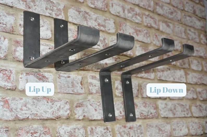 May include: Metal shelf brackets in a dark gray finish are mounted on a brick wall. The brackets are labeled "Lip Up" and "Lip Down". The brackets are designed to hold shelves and have a minimalist, industrial style.