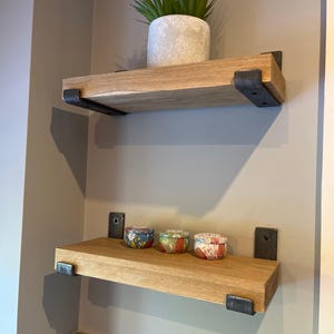 May include: Two rectangular wooden shelves with natural grain patterns, each mounted to the wall with dark metal brackets. The top shelf holds a potted green plant in a textured light grey container, while the bottom shelf holds three small round tins.