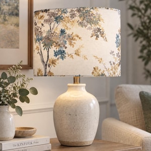 May include: A table lamp with a speckled cream ceramic base and a cylindrical fabric lampshade featuring a printed pattern of blue and yellow tree branches with leaves. The lamp has a metallic gold-toned neck connecting the base to the shade.