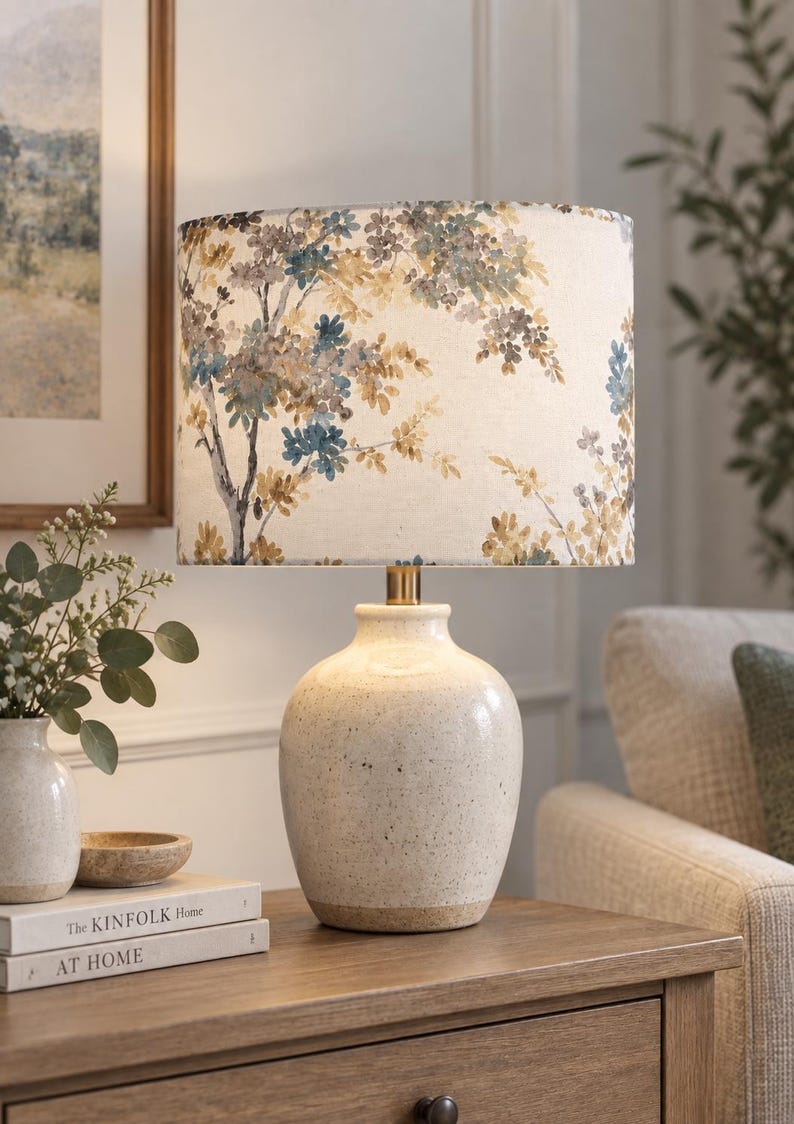 May include: A table lamp with a speckled cream ceramic base and a cylindrical fabric lampshade featuring a printed pattern of blue and yellow tree branches with leaves. The lamp has a metallic gold-toned neck connecting the base to the shade.